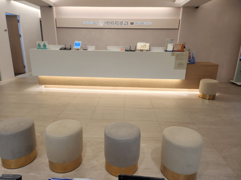 베이지 타일 바닥에 금색 밑단을 두른 원형 스툴 네 개가 앞쪽에 놓여 있고, 뒤로 흰색 리셉션 데스크와 '비수술 윤곽 성형 에버피부과 닥터필러 Dr. filler' 글자가 붙은 벽면 사인이 보이는 구도