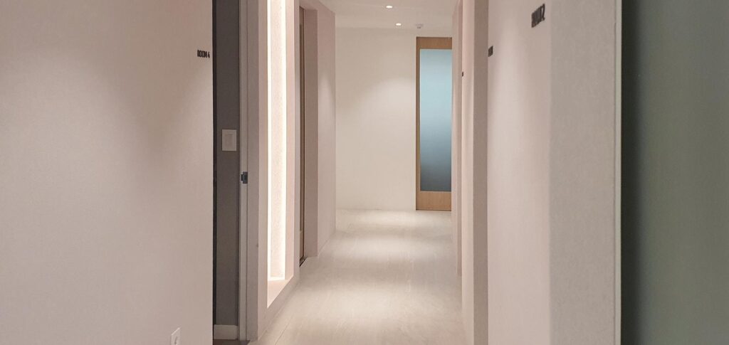 밝은 베이지 타일 바닥의 복도로, 왼쪽 벽에 ROOM 4 사인과 세로 간접조명이 있고 정면 끝에는 우드 프레임의 반투명 유리문이 보인다.