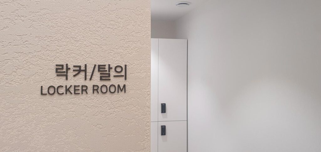 질감이 있는 베이지 벽면에 '락커/탈의 LOCKER ROOM' 입체 글자가 붙어 있고, 오른쪽으로 번호키가 달린 흰색 사물함이 세로로 놓여 있다.