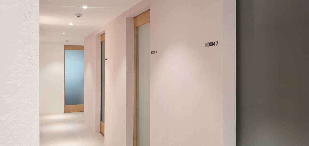 벽면에 'ROOM 2', 'ROOM 3' 글자가 붙어 있고 우드 프레임의 반투명 유리문이 이어지며, 복도 끝에는 파란빛이 도는 유리문이 보이는 가로 구도