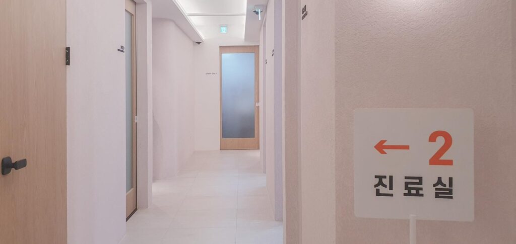 복도 벽면에 '← 2 진료실' 안내판이 걸려 있고, 왼쪽에는 우드 도어들이, 안쪽 끝에는 STAFF ONLY 표기와 반투명 유리문이 있다.