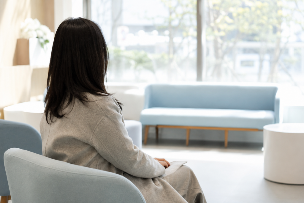 베이지색 코트를 입은 여성이 하늘색 의자에 앉은 뒷모습으로, 맞은편에 하늘색 벤치 소파와 큰 창이 있고 창밖으로 나무가 보이는 밝은 실내