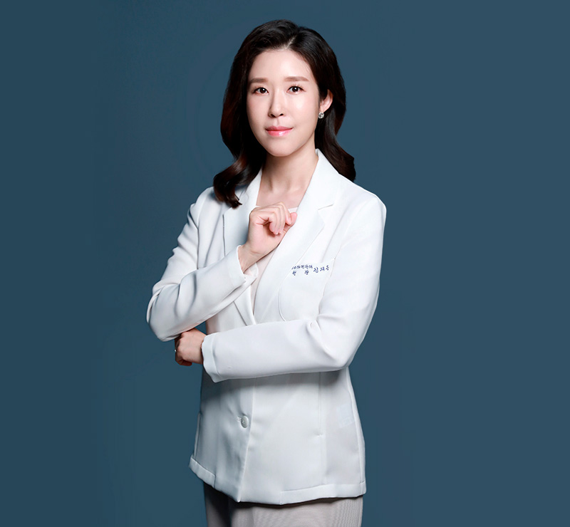짙은 청회색 배경 앞에서 흰색 가운을 입은 여성이 한 손을 가슴 앞에 모으고 정면을 바라보는 상반신 인물 사진